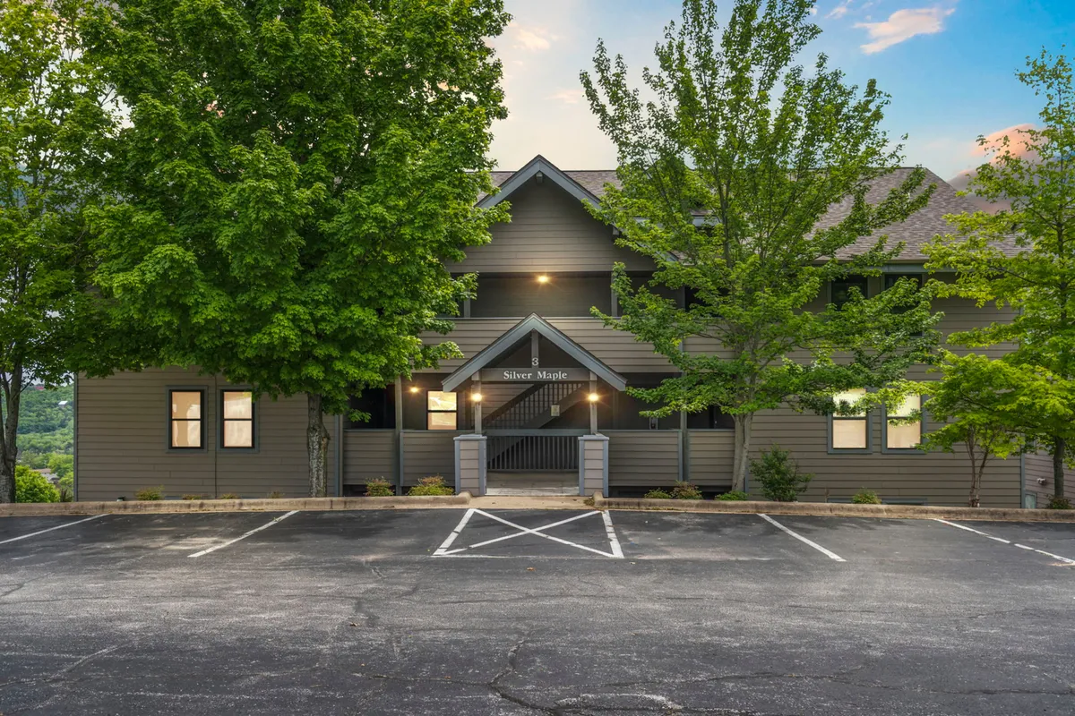 The image shows the building facade of a lakeside condo with a welcoming entrance and lush greenery surrounding the porch. The porch area is set up for relaxation, highlighted by seating that invites guests to enjoy the natural views and tranquil ambiance. The sunset lighting gives the exterior a romantic and cozy feel, perfect for morning coffee or evening chats.