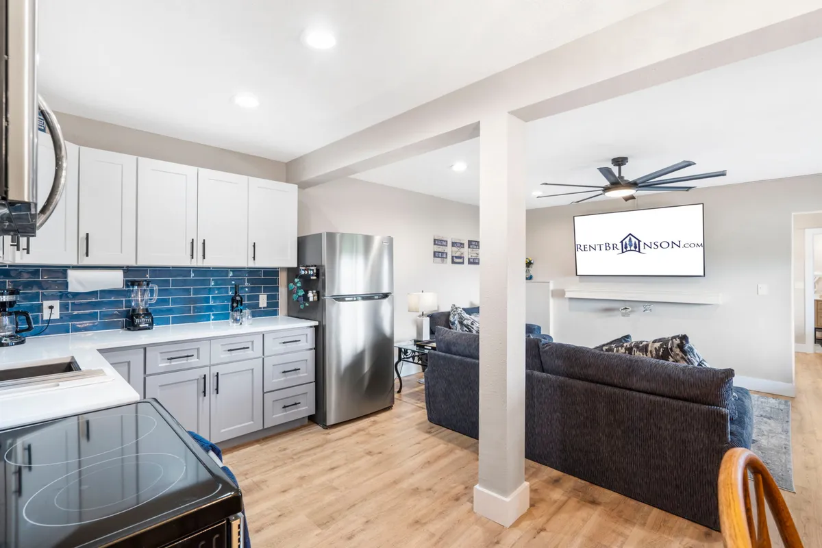 A cozy and stylish interior scene featuring a comfortable living room seating area arranged around a modern television, with the adjoining kitchen and its blue tile backsplash visible. The overall vibe is warm and inviting, ideal for both relaxing and socializing with loved ones.