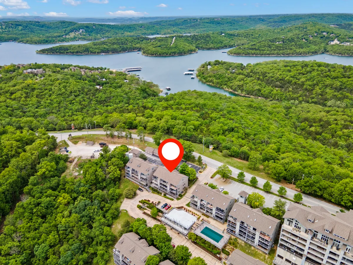 A waterfront view featuring lush greenery, residential buildings, and a sparkling swimming pool. The scene showcases opportunities for boating, hiking, and relaxing by the water, with a peaceful mood set by the serene landscape and open sky. The pool and balconies offer spaces to unwind and savor the scenic beauty of Table Rock Lake.