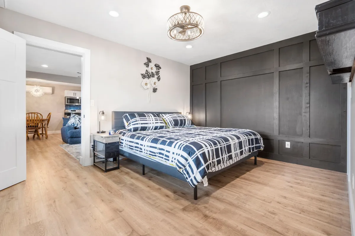 This image features a cozy bed with patterned bedding, a dark accent wall, a chandelier light fixture, and a doorway leading to another room. Natural light softly illuminates the space, creating a welcoming environment perfect for relaxation.