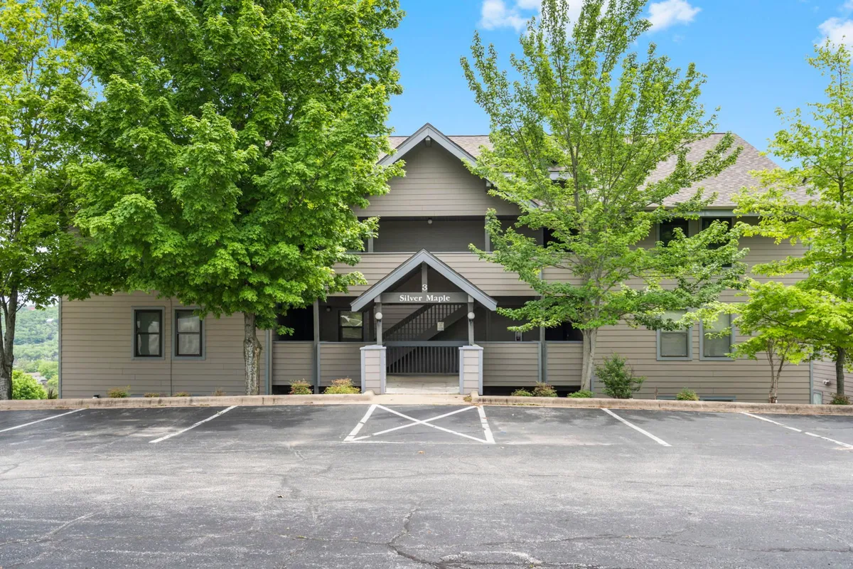 The image showcases the welcoming entrance to the Silver Maple condo building, framed by mature trees and a spacious parking area. The signage is clearly visible, and the setting exudes a serene, inviting vibe that hints at group gatherings and easy arrivals.
