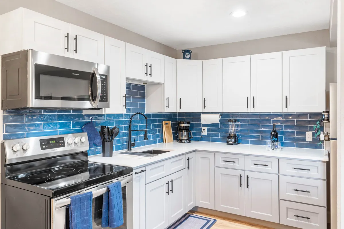 The image showcases a sparkling, freshly remodeled kitchen with gleaming stainless steel appliances, a striking blue tile backsplash, and crisp white cabinetry. The scene feels open and welcoming, inviting guests to cook, gather, and enjoy time together. The kitchen's modern design is both stylish and functional, ideal for both relaxed breakfasts and special evening meals.