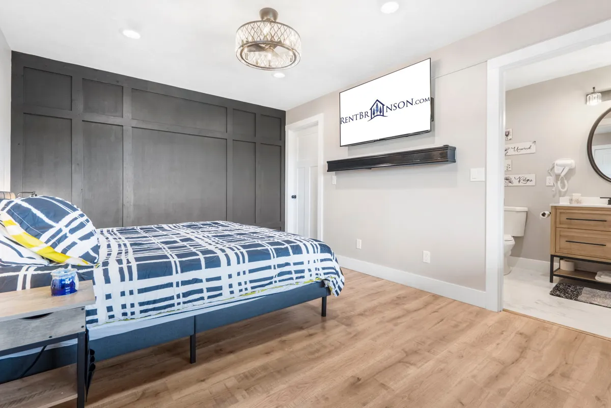 The photo depicts a comfortable bedroom with patterned bedding, a wall-mounted TV, a dark accent wall, and a nightstand adorned with a decorative item. The room exudes a modern yet restful vibe, emphasizing comfort and style.